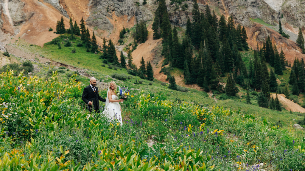 KatieToddPhotography 2 - Beaumont Hotel Wedding couple in formal attire walking through a lush wildflower meadow with a mountainous backdrop and evergreen trees behind them.