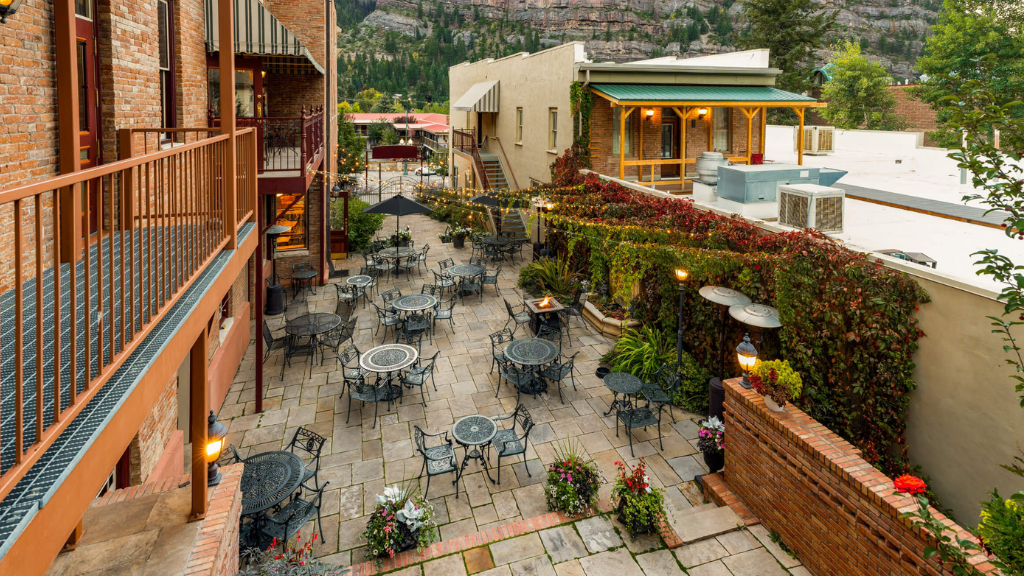 Brick-walled outdoor patio with metal tables and chairs, string lights, and potted plants in a courtyard setting.