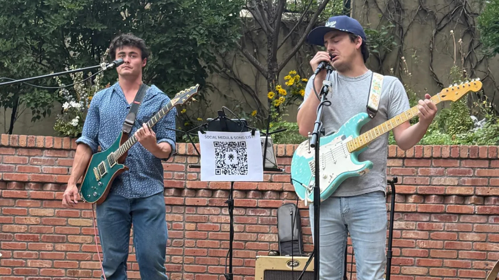 Two men perform on a brick stage: left guitarist in a blue patterned shirt with a teal guitar, right singer with a mint Stratocaster and a microphone, as a QR code sign for social media and song list sits between them.