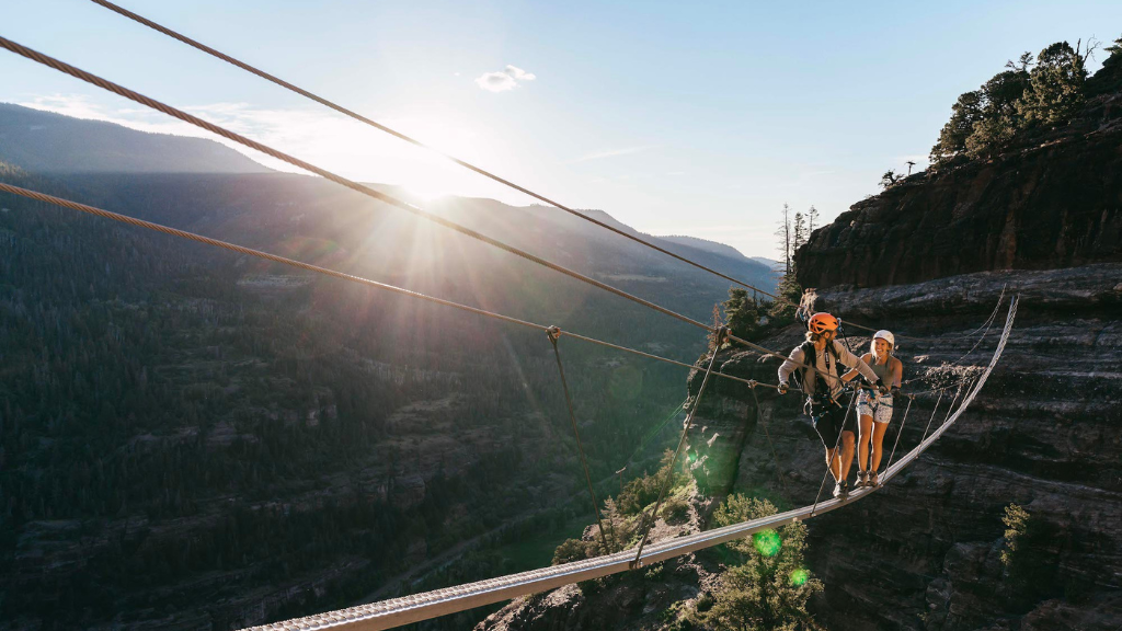 Two hikers wearing helmets cross a narrow suspension bridge between cliff faces at sunset.