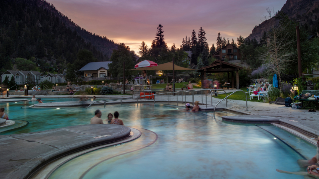 People soak in a winding outdoor pool at a mountain resort during sunset with trees and buildings in the background.