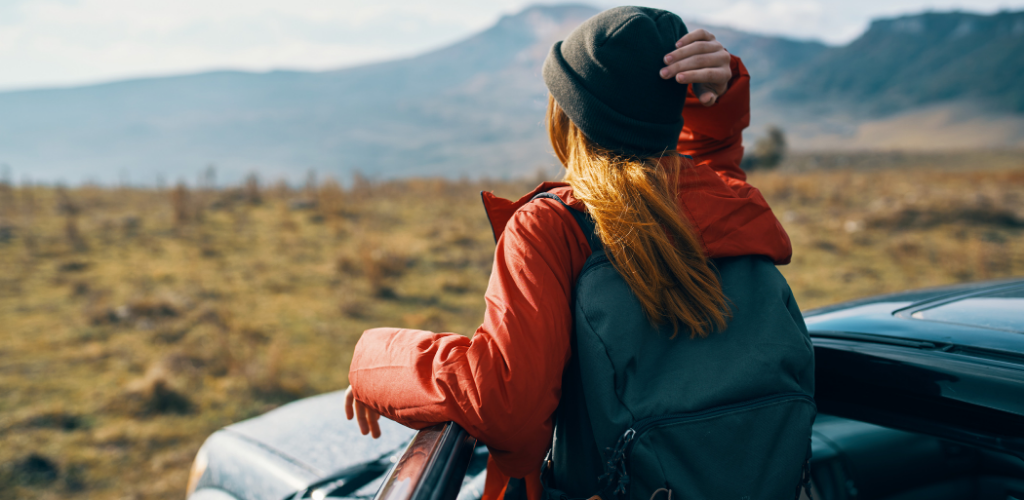 2 - Beaumont Hotel Person with a backpack leaning on an SUV in a open veld, gazing at distant mountains; wearing orange jacket and beanie in daytime light.
