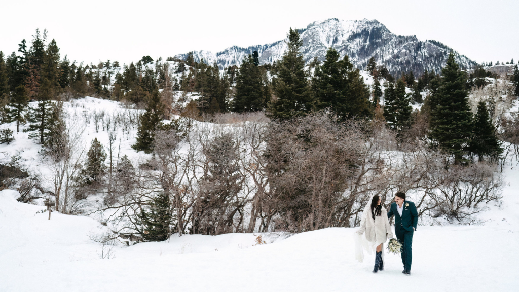 Winter wedding - Beaumont Hotel A bride and groom walk through a snowy landscape with evergreen trees and mountains in the background.